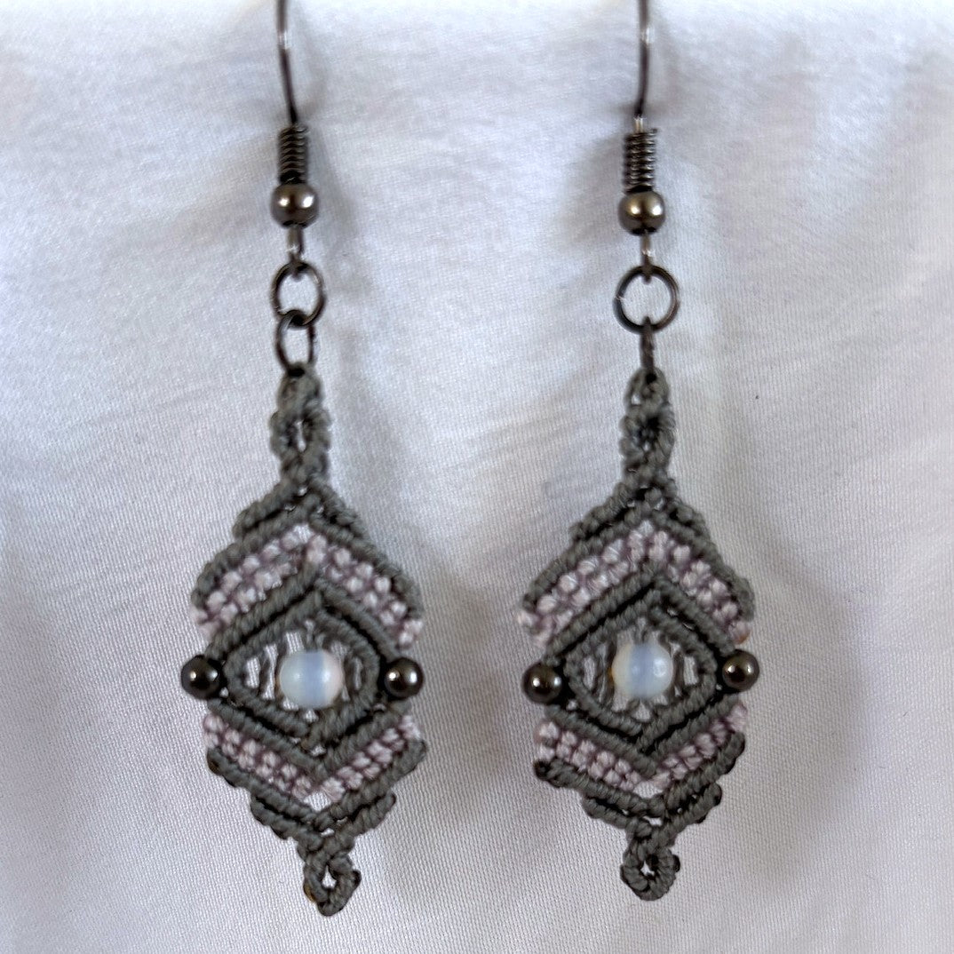 Pair of handmade medium silvery gray nylon macrame drop earrings with open-center diamond silhouette, pink cord accent rows, 4mm round opalite center bead, 3mm gunmetal round metal bead accents, and gunmetal fish hook wires, displayed on white fabric background.