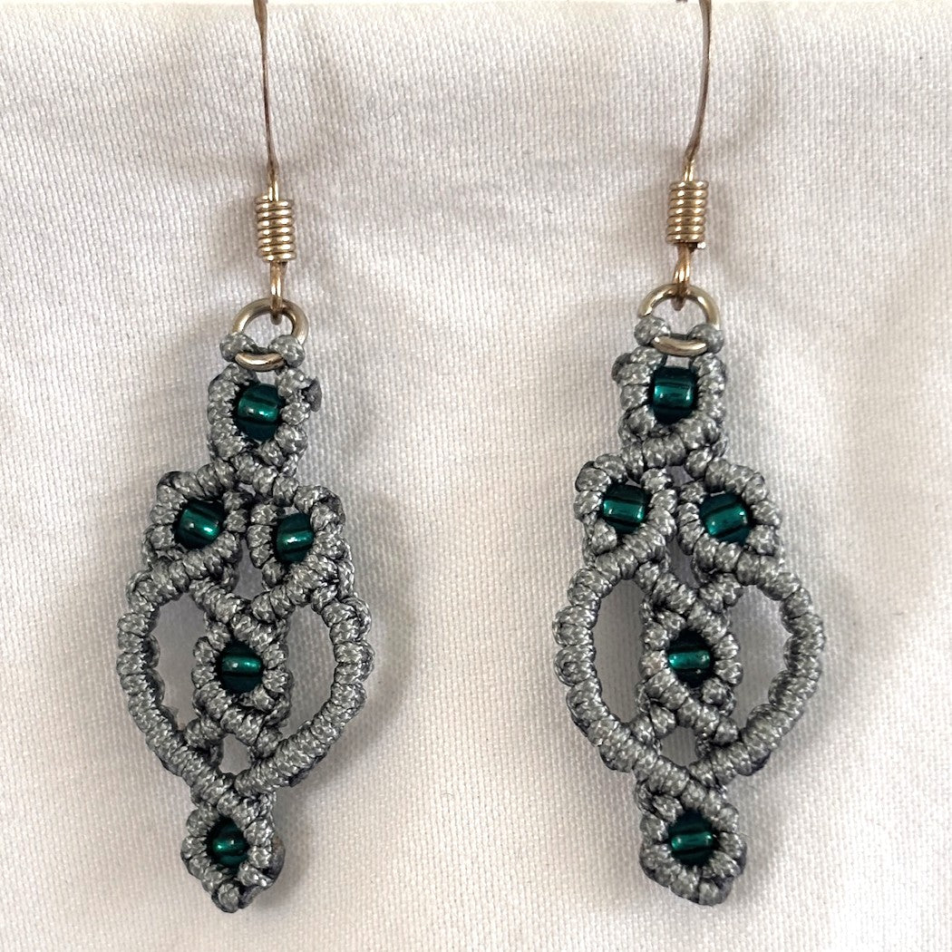 Pair of handmade gray nylon macrame drop earrings with interlocking ring and picot lattice knotwork, 8/0 emerald green seed beads, and silver-tone fish hook wires, displayed on white fabric background.