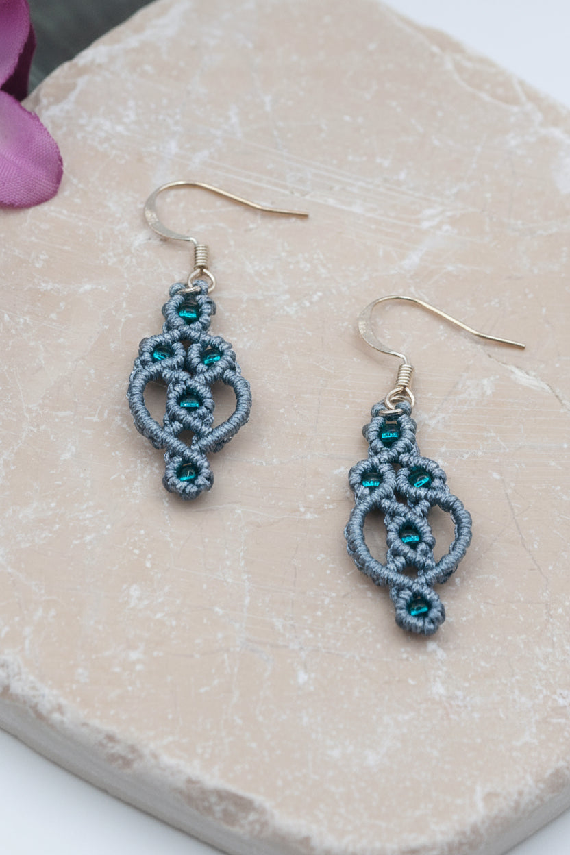 Handmade gray nylon macrame drop earrings with interlocking rings and picots, emerald green seed beads woven throughout, and silver-tone fish hook wires, displayed on a natural stone surface with a pink flower.
