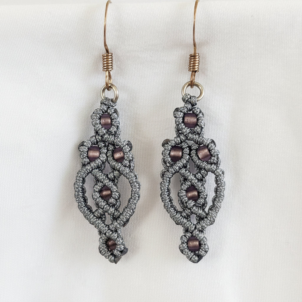 Pair of handmade gunmetal gray nylon macrame lace drop earrings with elongated diamond lattice silhouette, 8/0 plum seed bead accents, and silver-tone fish hook wires, displayed on white fabric background.