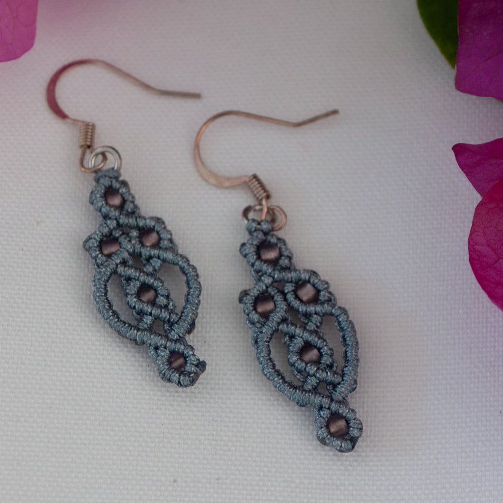 Handmade gunmetal gray nylon macrame lace drop earrings with an elongated diamond lattice silhouette, plum seed beads woven throughout, and silver-tone fish hook wires, displayed on a white surface with magenta bougainvillea flowers.