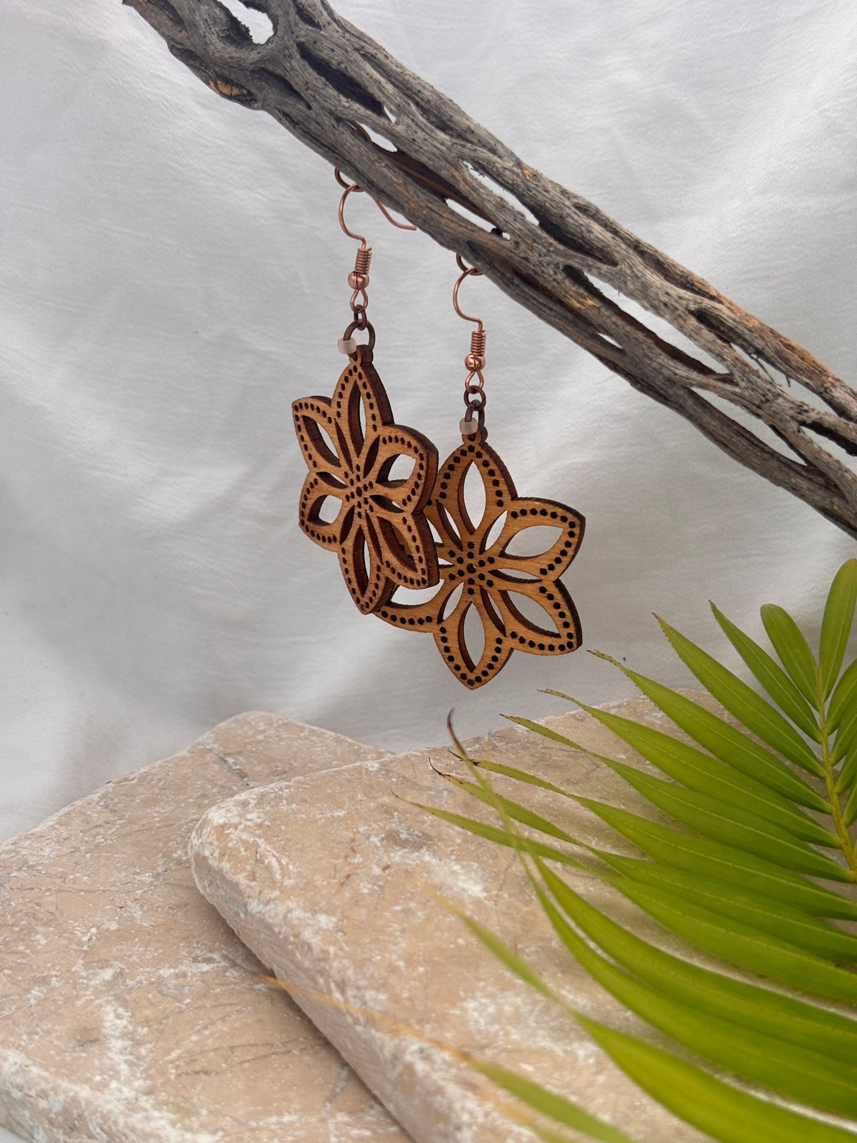 Handmade balsa wood flower earrings with open petal silhouette and dotted border detail with frosted peach seed beads hanging from a driftwood branch with a stone and palm leaf backdrop.