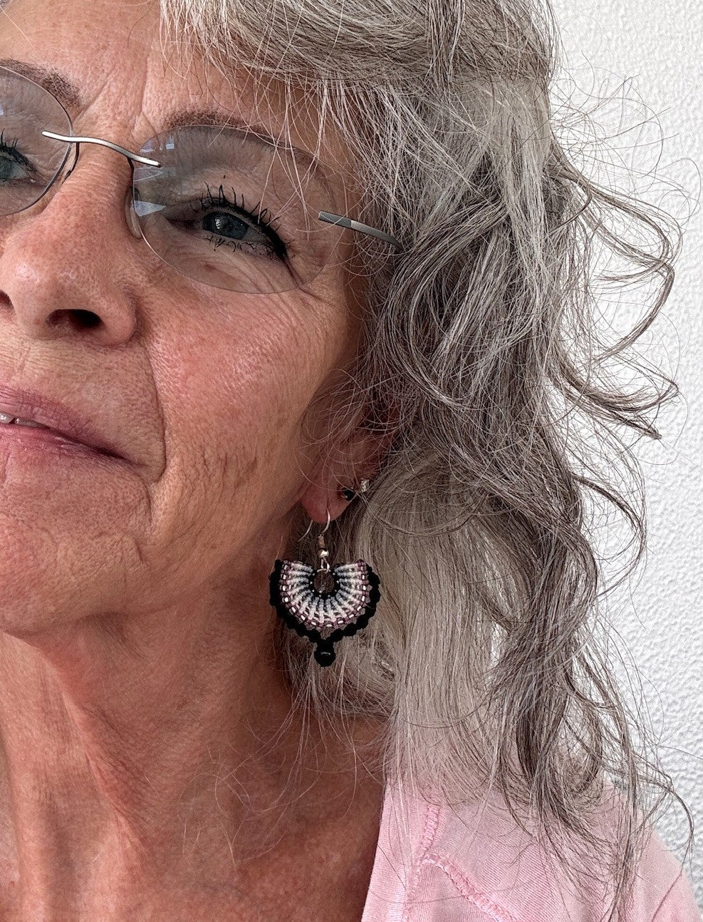 Handmade black macrame fan drop earring with gradient knotwork in black, steel gray, ivory, pink, and mauve cord, size 11/0 silver-lined seed bead accents, size 8/0 amethyst seed bead drop, and silver-tone fishhook ear wire, shown worn on model with silver curly hair and rimless glasses.