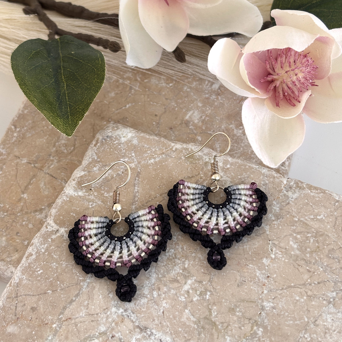 Pair of handmade black macrame fan drop earrings with gradient knotwork in black, steel gray, ivory, pink, and mauve cord, size 11/0 silver-lined seed bead accent rows, size 8/0 amethyst seed bead macrame loop drop, and silver-tone fish hook wires, displayed on travertine stone with magnolia flower and green leaf.
