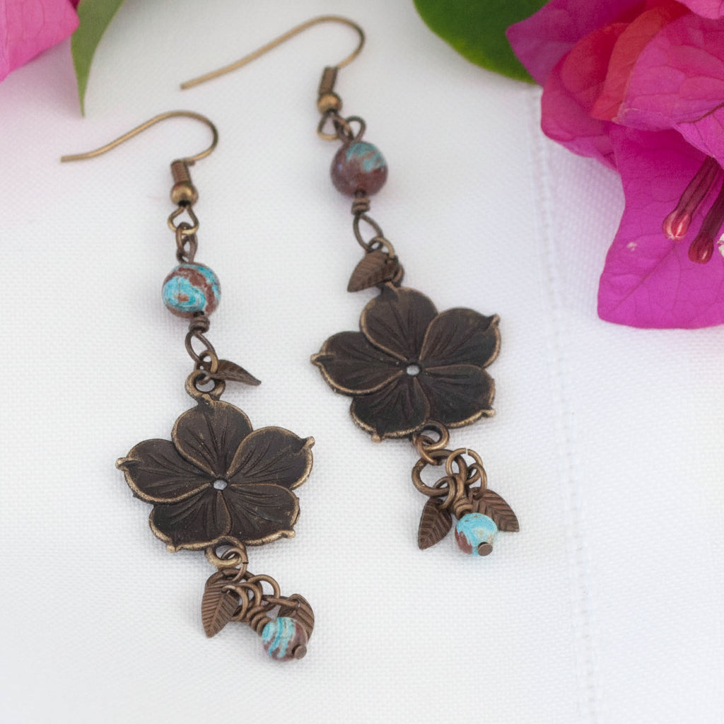 Pair of bronze flower charm earrings with calsilica beads and leaf accent drops, flat lay on white surface with pink bougainvillea