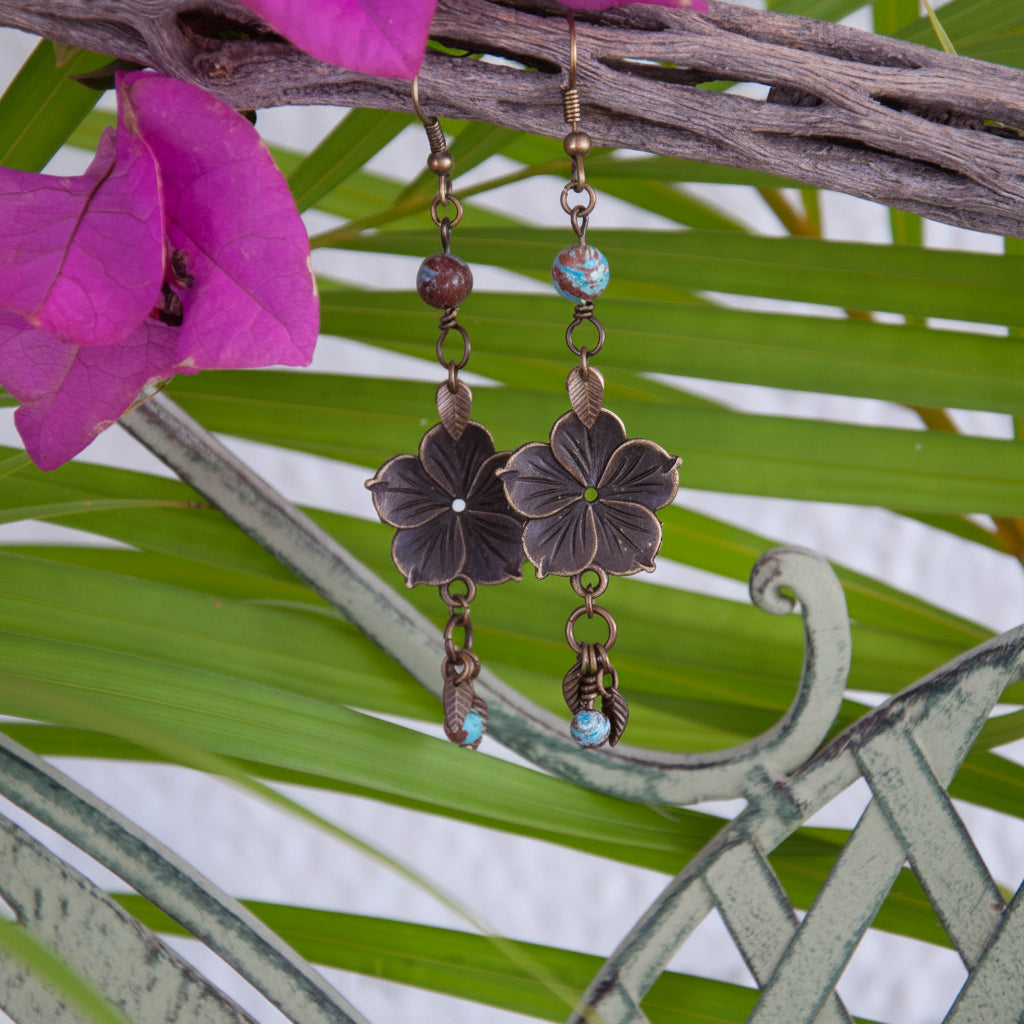 Bronze hibiscus flower charm earrings with teal and brown calsilica beads, hanging from driftwood with bougainvillea and tropical palm frond