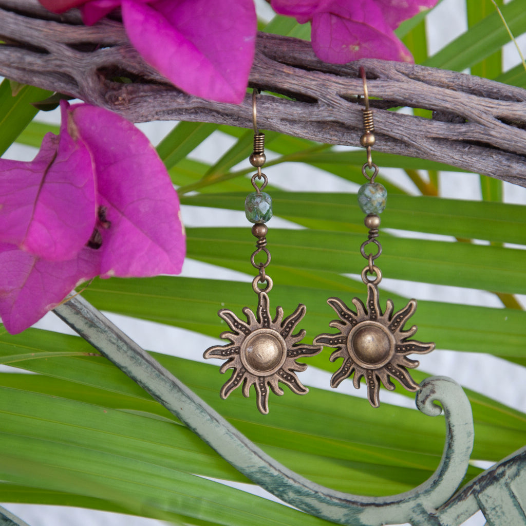 Bronze sunburst charm earrings with green Picasso Czech beads, hanging from driftwood with tropical palm leaves and bougainvillea