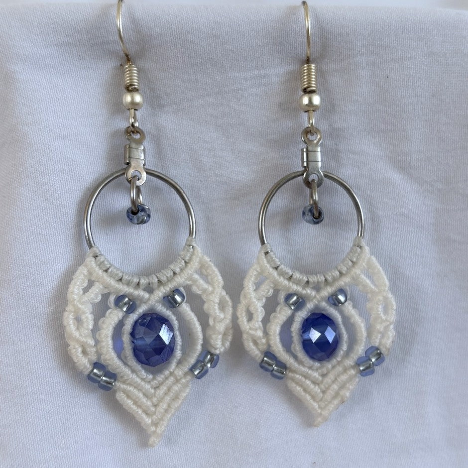 Pair of handmade white macrame fan hoop drop earrings with 16mm silver-tone hoop, 6mm × 8mm faceted blue crystal rondelle center bead, size 8/0 silver-lined light blue seed beads, and silver-tone fish hook wires, displayed on white fabric background.