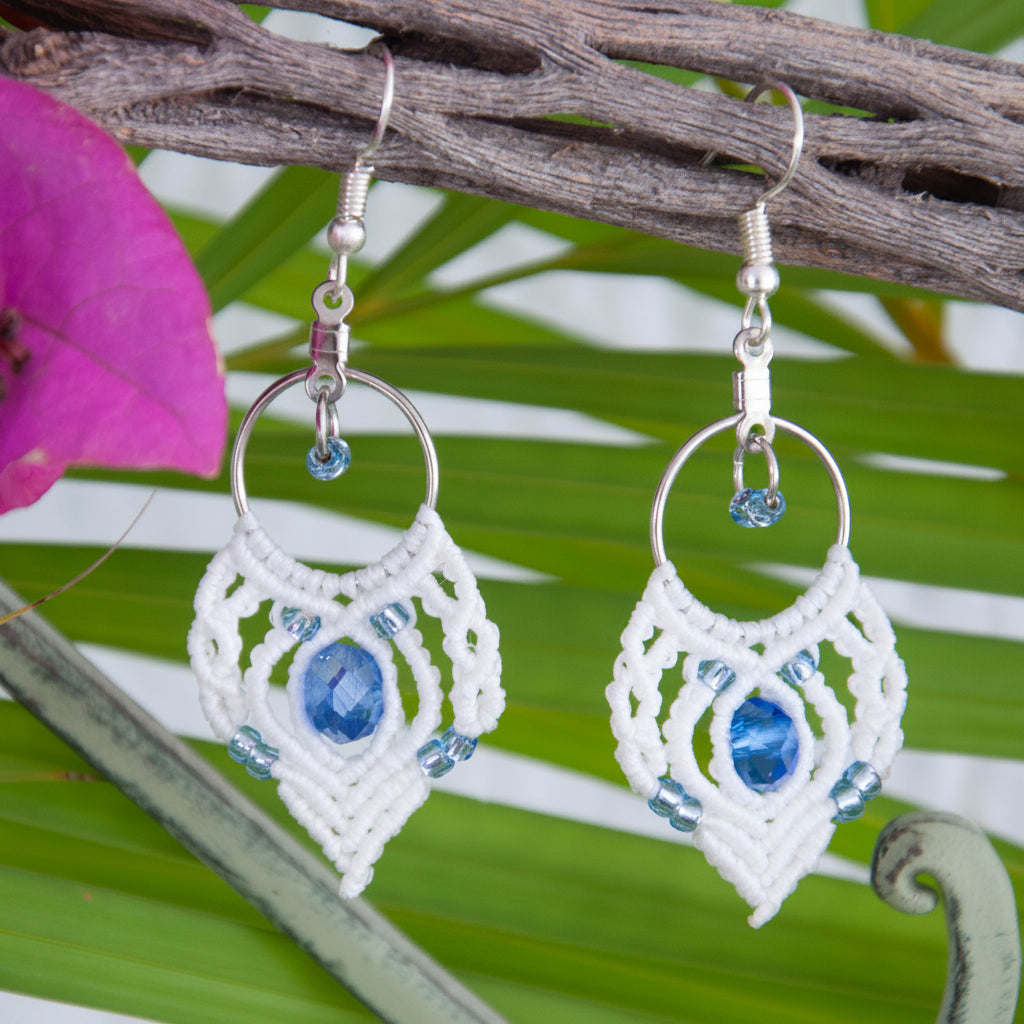 Handmade white macrame fan hoop drop earrings with a silver-tone hoop finding, faceted blue crystal rondelle center bead, silver-lined light blue seed beads, and silver-tone fish hook wires, hanging from a driftwood branch with a tropical palm leaf and magenta bougainvillea flowers.