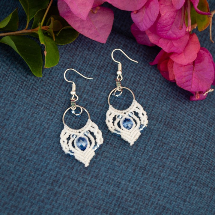 Handmade white macrame fan hoop drop earrings with a silver-tone hoop finding, faceted blue crystal rondelle center bead, silver-lined light blue seed beads, and silver-tone fish hook wires, laid on a dark blue-gray textured surface with magenta bougainvillea flowers.