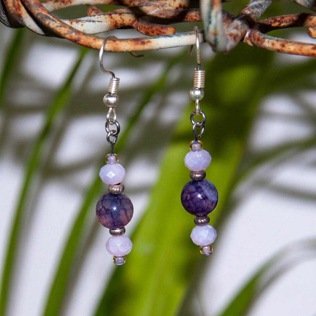 Handmade amethyst and lavender crystal drop earrings with faceted lavender rondelle crystal beads, lavender seed bead accents, textured silver-tone spacer beads, and silver-tone fishhook ear wires, hanging from a rustic wire branch with tropical green leaves.