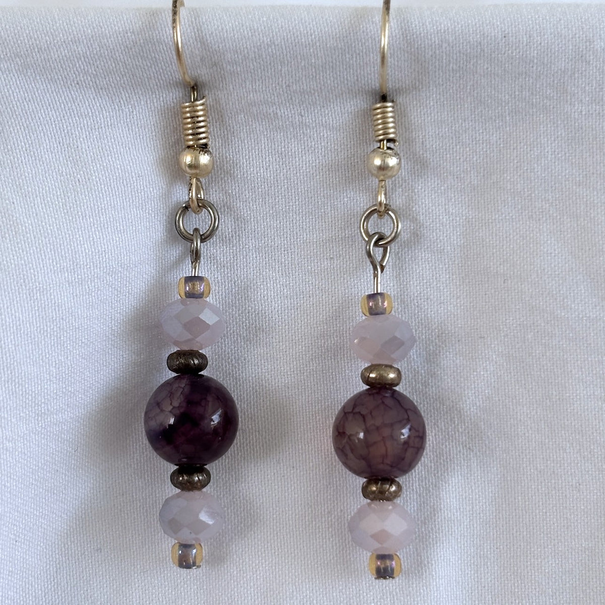 Pair of handmade amethyst and lavender crystal drop earrings with 8mm round amethyst beads, 4mm × 6mm faceted lavender rondelle crystal beads above and below, size 8/0 lavender seed bead accents, textured silver-tone spacer beads, and silver-tone fishhook ear wires, displayed on white fabric background.