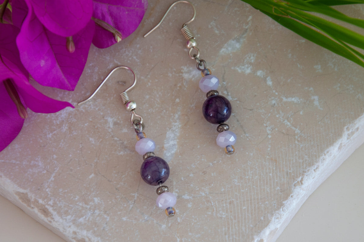 Handmade amethyst and lavender crystal drop earrings with faceted lavender rondelle crystal beads, lavender seed bead accents, textured silver-tone spacer beads, and silver-tone fishhook ear wires, laid on travertine tile with magenta bougainvillea and tropical palm leaves.