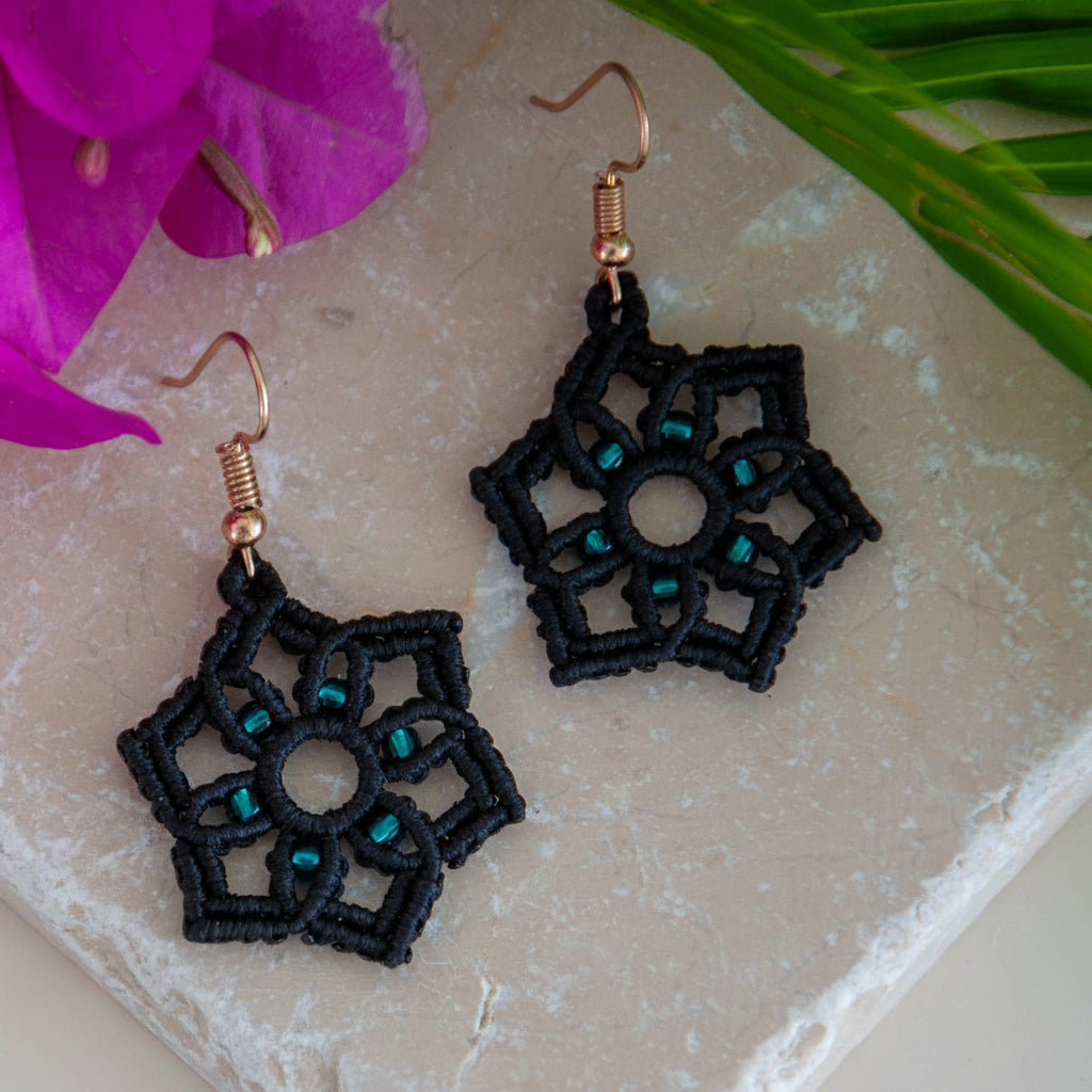 Handmade black macrame star drop earrings with silver-lined emerald green seed bead accents and gold-tone fish hook wires, laid on a travertine tile with a magenta orchid and tropical palm leaves.