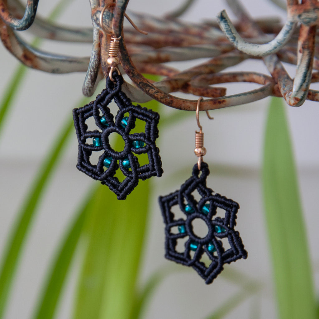 Handmade black macrame star drop earrings with silver-lined emerald green seed bead accents and gold-tone fish hook wires, hanging from a rustic metal stand with tropical green leaves in the background.