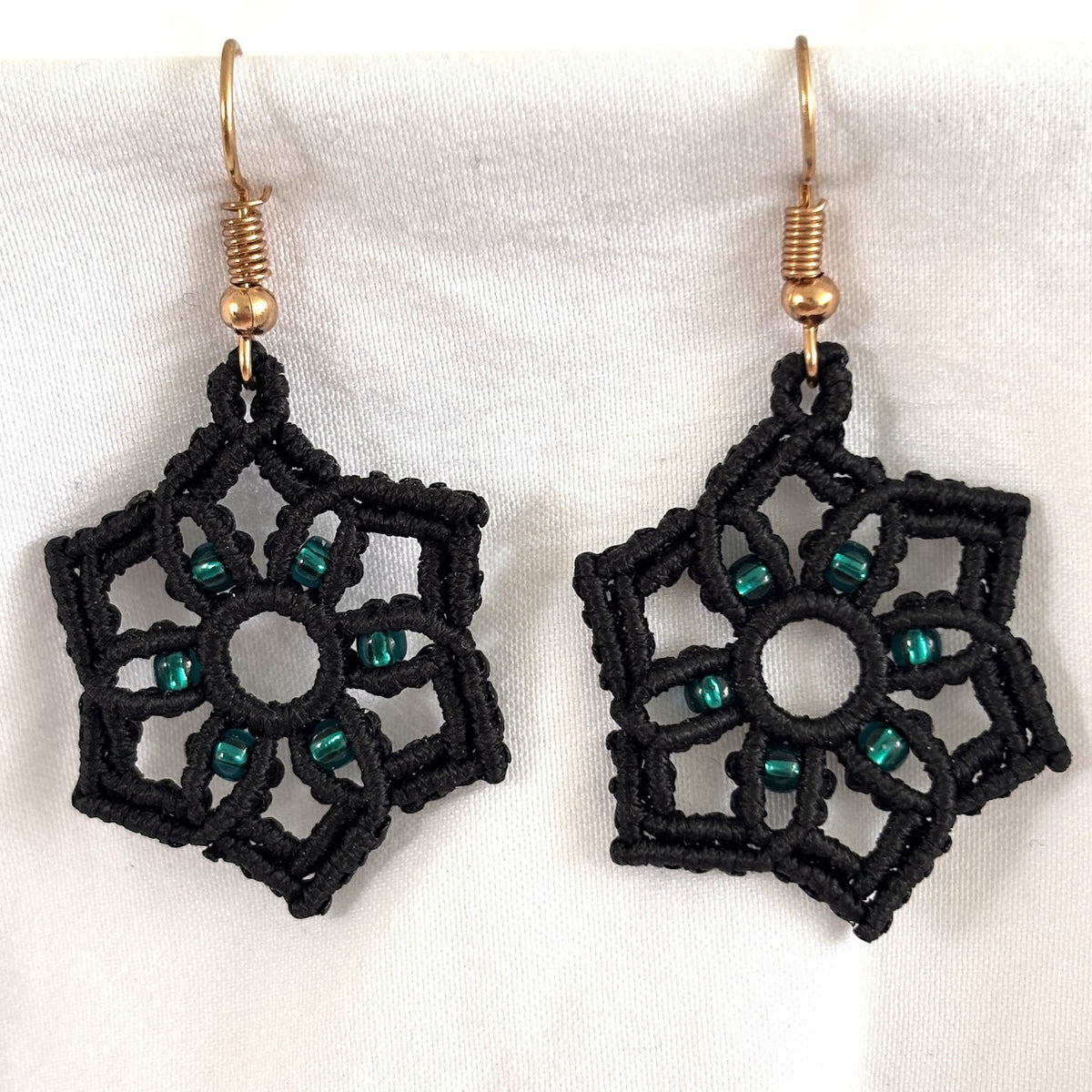 Pair of handmade black macrame star drop earrings with 8/0 silver-lined emerald green seed bead accents at each point, open circle center, and gold-tone fish hook wires, displayed on white fabric background.