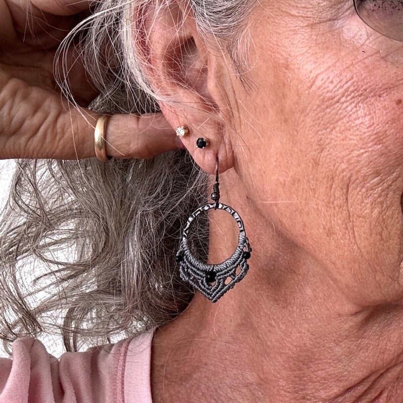 Handmade macrame hoop earring with 25mm hammered gunmetal ring, gray macrame lattice knotwork, 4mm round black onyx center bead, 2mm round black metal accent beads, and gunmetal fishhook ear wire, shown worn on model with silver hair.
