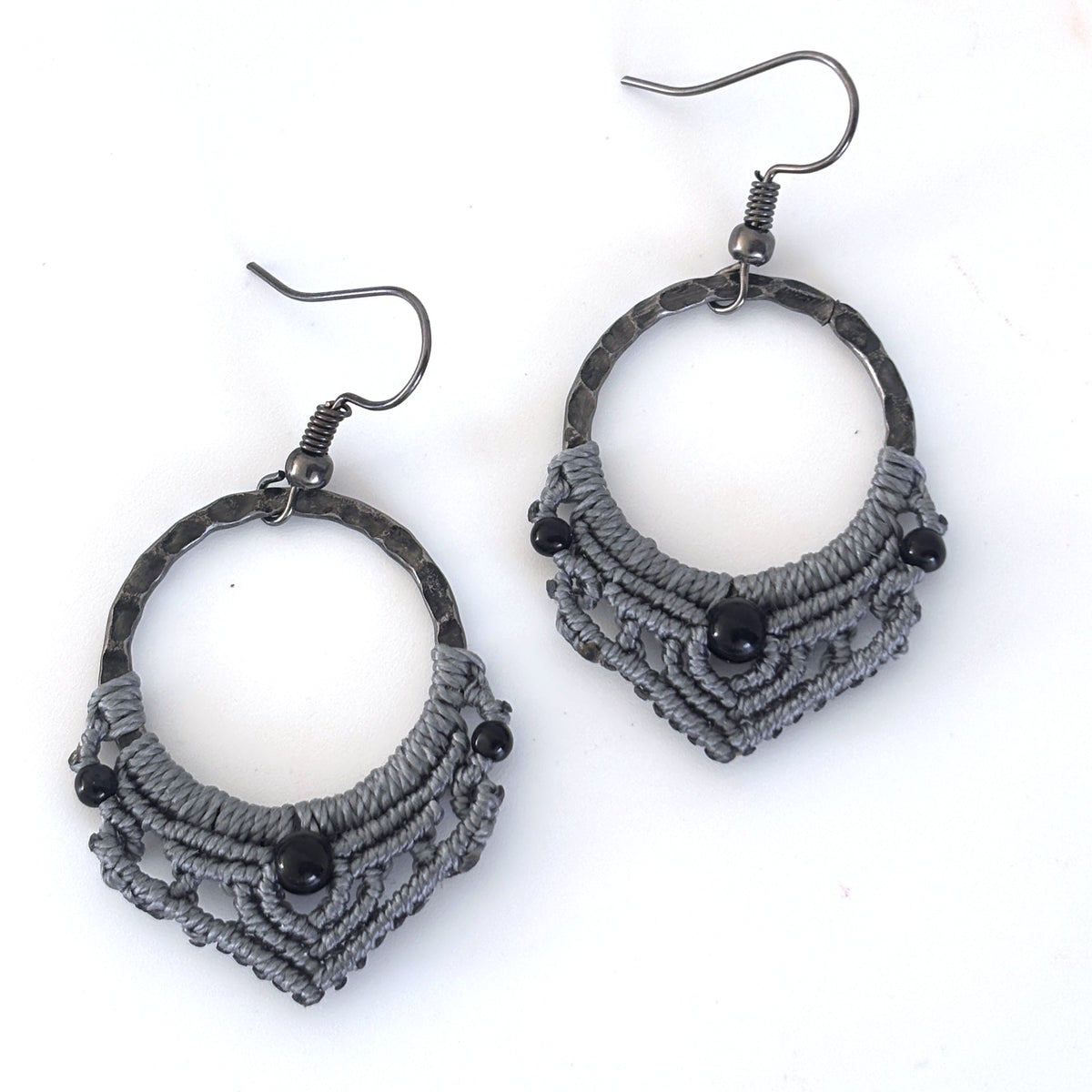 Pair of handmade macrame hoop earrings with 25mm hammered gunmetal rings, gray macrame lattice knotwork, 4mm round black onyx center beads, 2mm round black metal accent beads, and gunmetal fishhook ear wires, displayed on white background showing full construction and bead detail.
