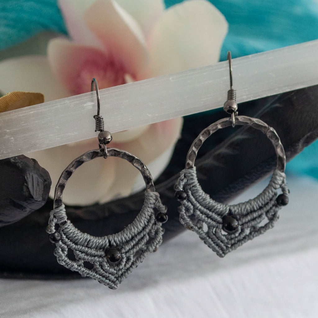 Pair of handmade macrame hoop earrings with 25mm hammered gunmetal rings, gray macrame lattice knotwork, 4mm round black onyx center beads, 2mm round black metal accent beads, and gunmetal fishhook ear wires, resting on black stone with selenite wand and magnolia flower against teal backdrop.
