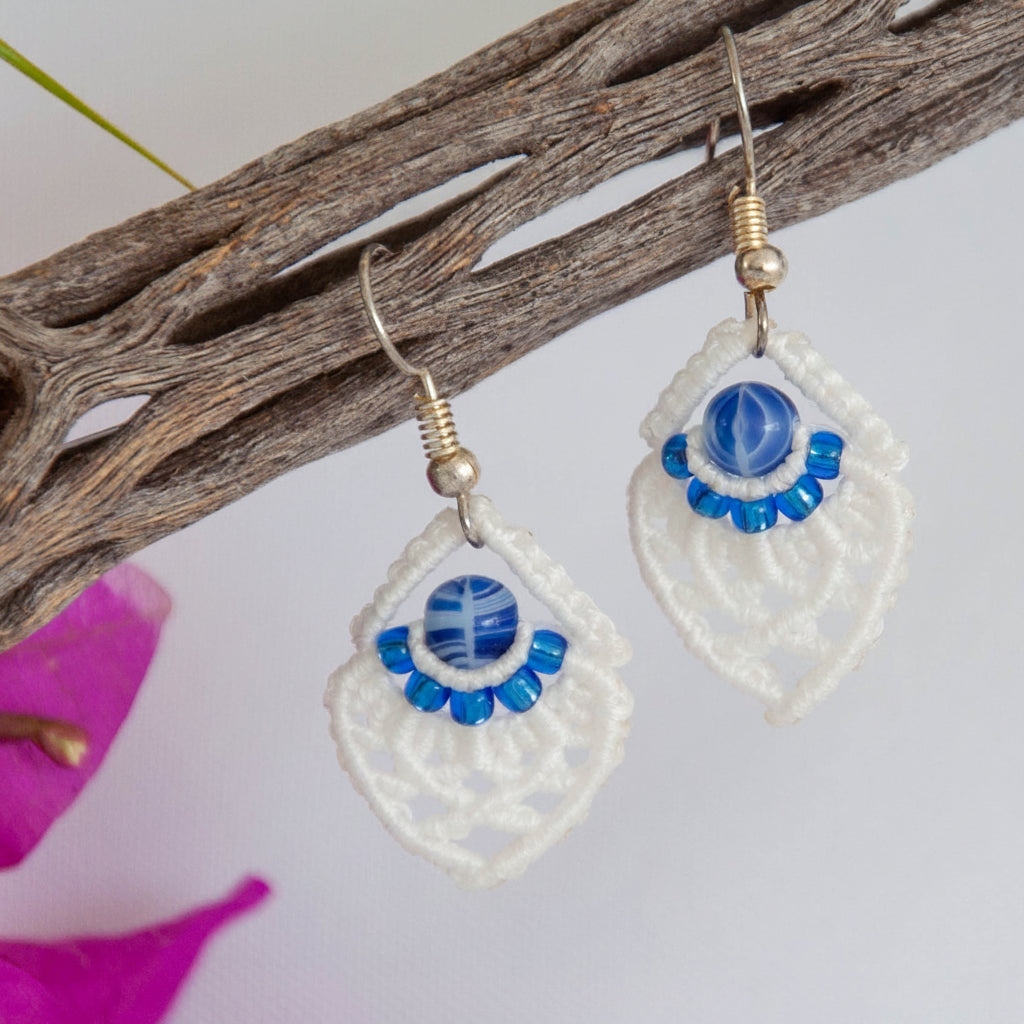 Handmade white macrame diamond drop earrings with silver-lined sapphire blue seed beads, a blue and white marbled glass center bead, and silver-tone fish hook wires, hanging from a driftwood branch with magenta bougainvillea against a white background.