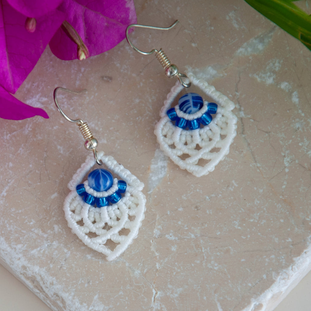 Handmade white macrame diamond drop earrings with silver-lined sapphire blue seed beads, a blue and white marbled glass center bead, and silver-tone fish hook wires, laid on a travertine tile with magenta bougainvillea and tropical green leaves.