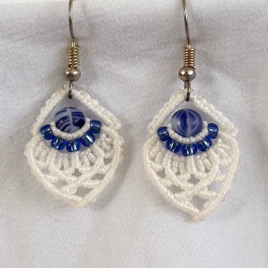 Pair of handmade white macrame diamond drop earrings with 8/0 silver-lined sapphire blue seed beads, 6mm blue and white marbled glass center beads, and silver-tone fish hook wires, displayed on white fabric background.