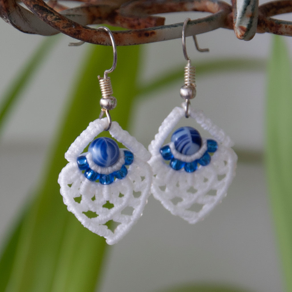 Handmade white macrame diamond drop earrings with silver-lined sapphire blue seed beads, a blue and white marbled glass center bead, and silver-tone fish hook wires, hanging from a rustic metal stand with tropical green leaves in the background.
