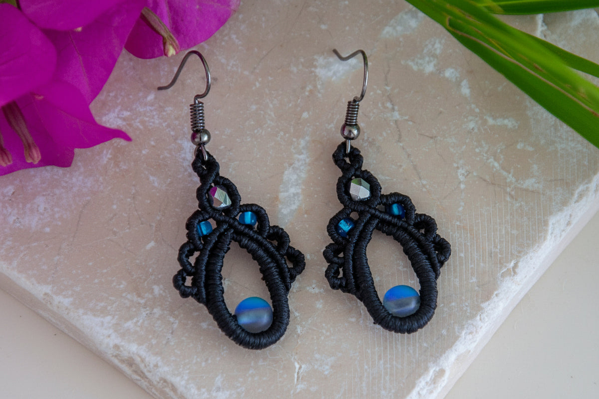 Handmade black macrame teardrop drop earrings with an iridescent gray mermaid glass center bead, silver-lined sapphire blue seed beads, faceted silver glass bead accent, and gunmetal fish hook wires, laid on a travertine tile with magenta bougainvillea and tropical palm leaves.