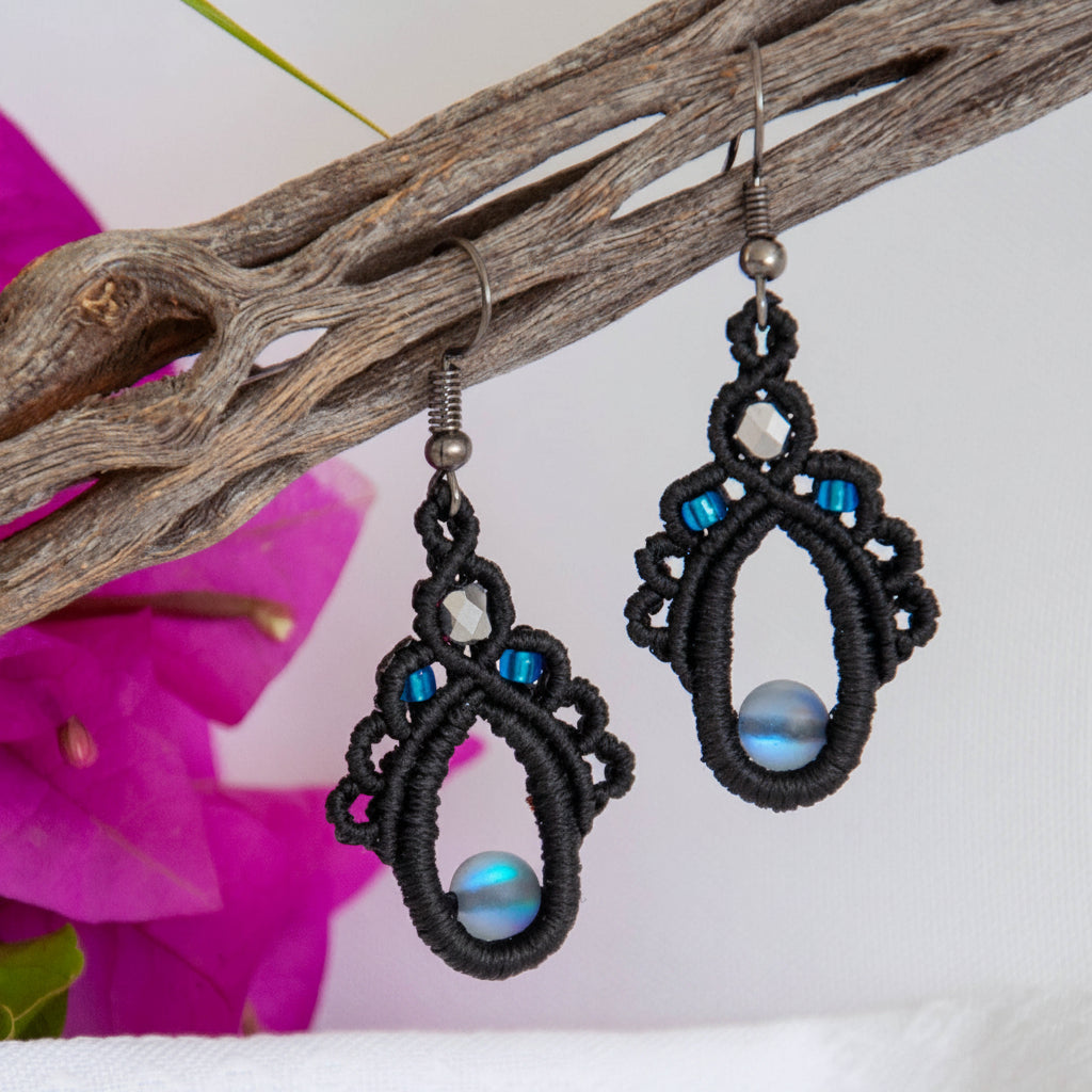 Handmade black macrame teardrop drop earrings with an iridescent gray mermaid glass center bead, silver-lined sapphire blue seed beads, faceted silver glass bead accent, and gunmetal fish hook wires, hanging from a driftwood branch with magenta bougainvillea against a white background.