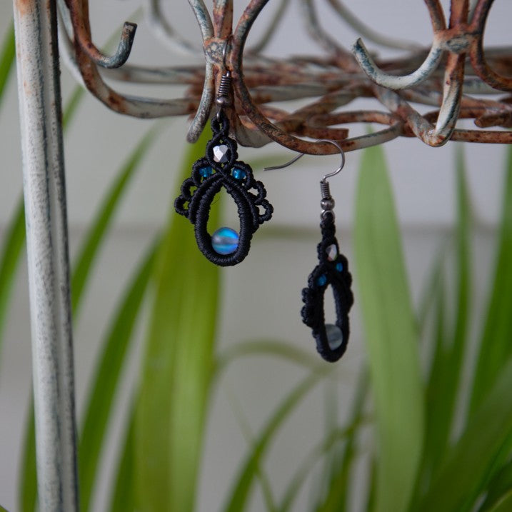 Handmade black macrame teardrop drop earrings with an iridescent gray mermaid glass center bead, silver-lined sapphire blue seed beads, faceted silver glass bead accent, and gunmetal fish hook wires, hanging from a rustic metal stand with tropical green leaves in the background.