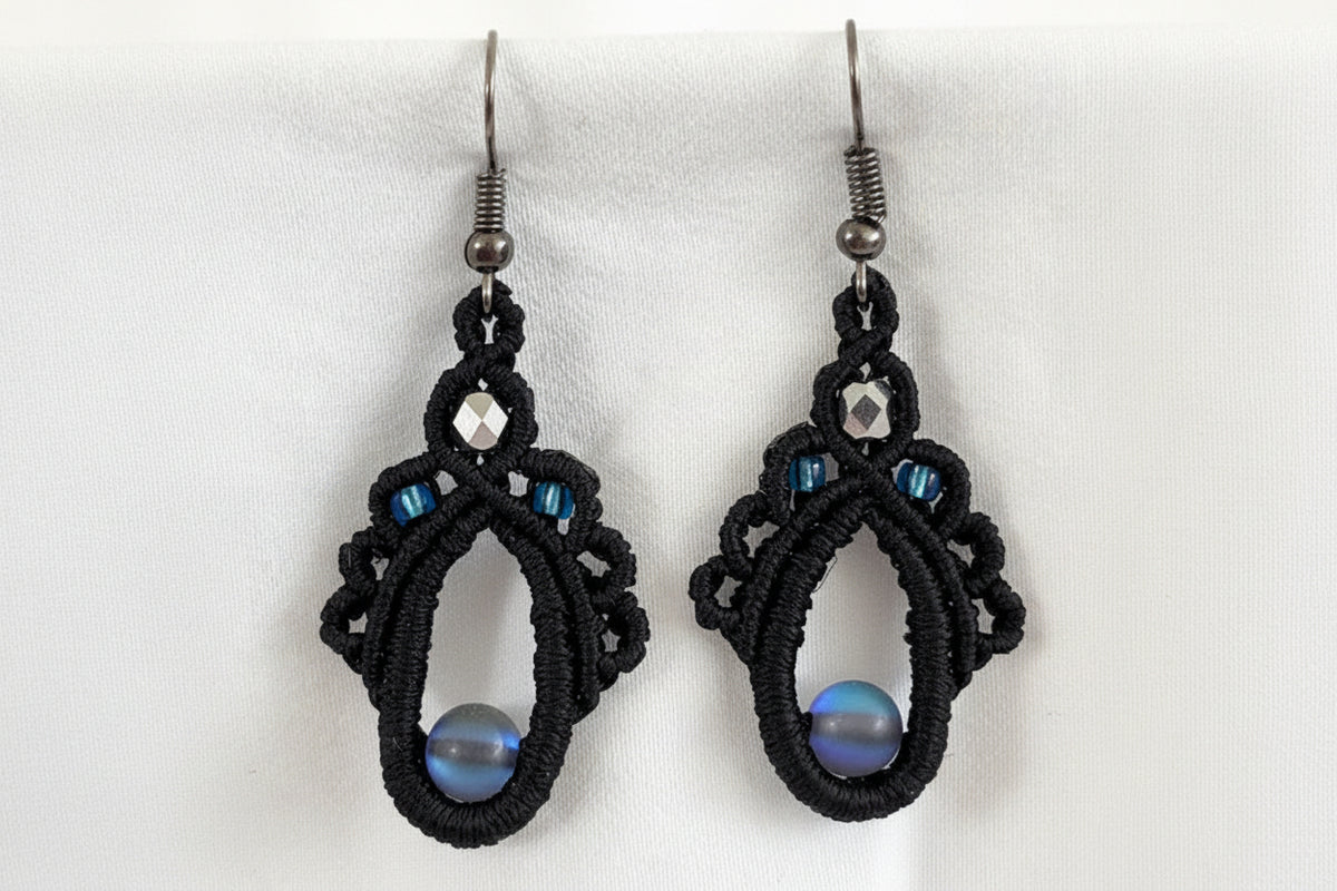 Pair of handmade black macrame teardrop drop earrings with 6mm iridescent gray mermaid glass center bead, 4mm faceted silver Czech glass crown bead, 8/0 silver-lined sapphire blue seed beads, and gunmetal fish hook wires, displayed on white background.