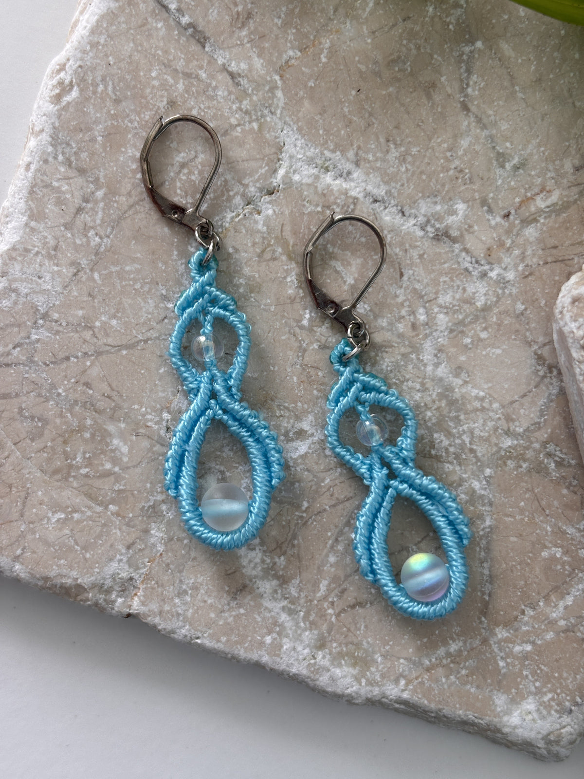 Pair of handmade turquoise macrame double drop earrings with iridescent mermaid glass beads and silver-tone lever-back closures, laid flat on a natural stone surface.