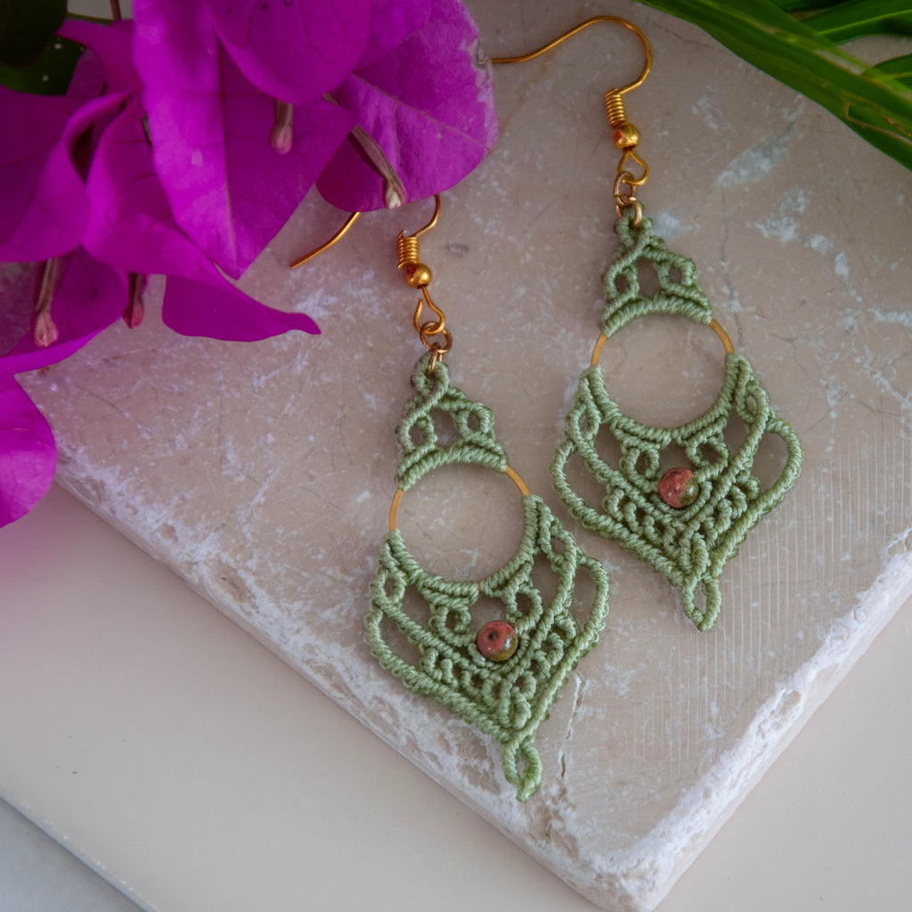 Handmade sage green lace macrame chandelier earrings with unakite bead and gold-tone ring, flat lay on travertine stone with bougainvillea.