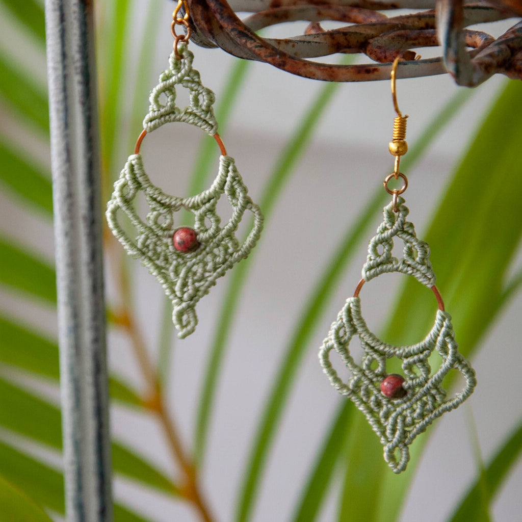 Pair of handmade sage green lace macrame chandelier earrings with unakite bead, displayed on rustic metal stand with tropical palm frond
