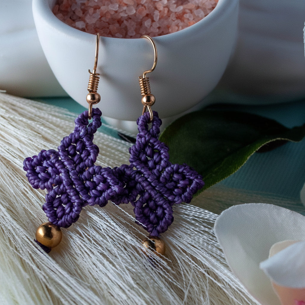 Handmade violet purple macrame four-leaf clover drop earrings with brass bead accents and gold-tone fish hook wires, laid on a white feather with tropical green leaves, a white ceramic bowl of Himalayan salt, and a white flower in the background.