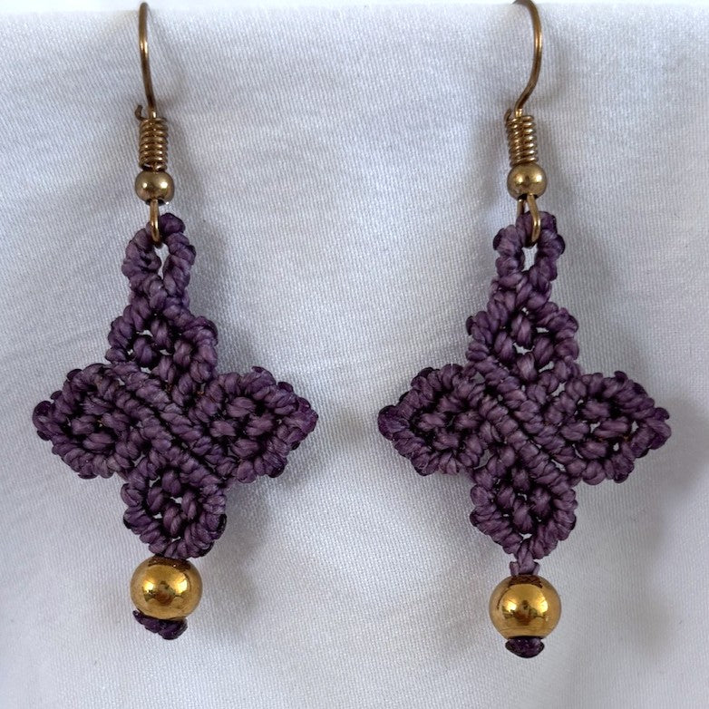 Pair of handmade violet purple Linhasita macrame four-leaf clover drop earrings with 6mm brass bead accents and gold-tone fish hook wires, displayed on white fabric background showing detailed knotwork.