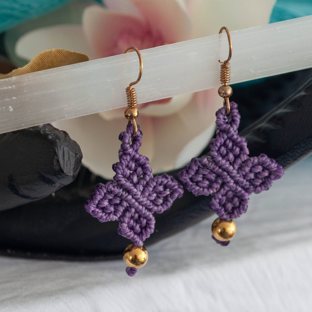 Handmade violet purple macrame four-leaf clover drop earrings with brass bead accents and gold-tone fish hook wires, resting on a white selenite wand over a black stone dish with a pink flower.