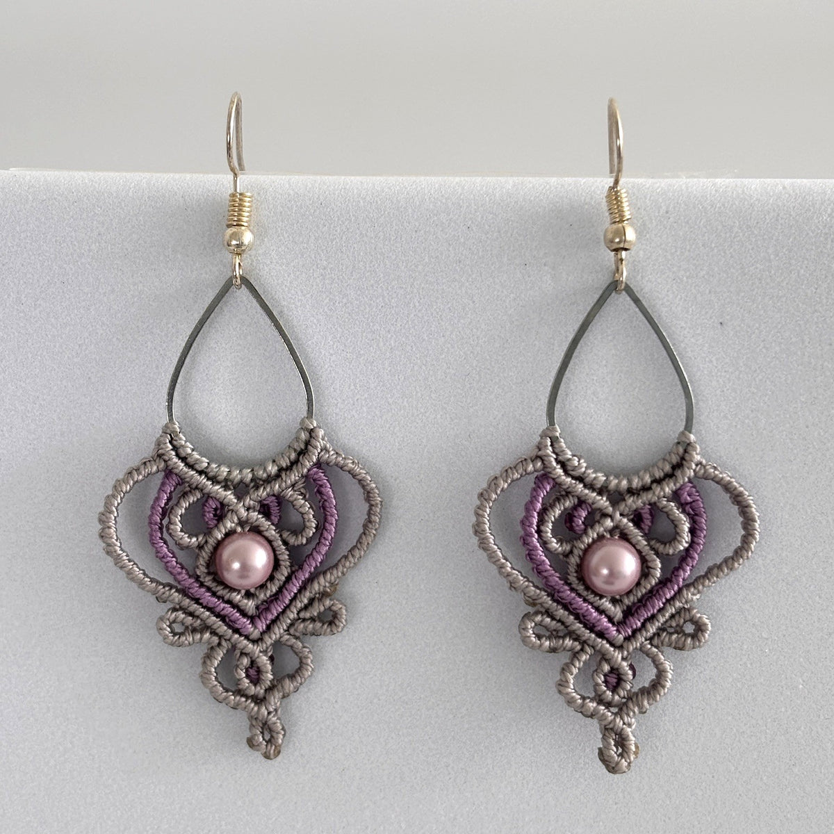 Close-up of handmade two-tone silvery gray and purple macrame teardrop drop earrings with ornate heart-shaped lattice, mauve shell pearl center bead, and silver-tone fish hook wires, displayed on a light gray surface.
