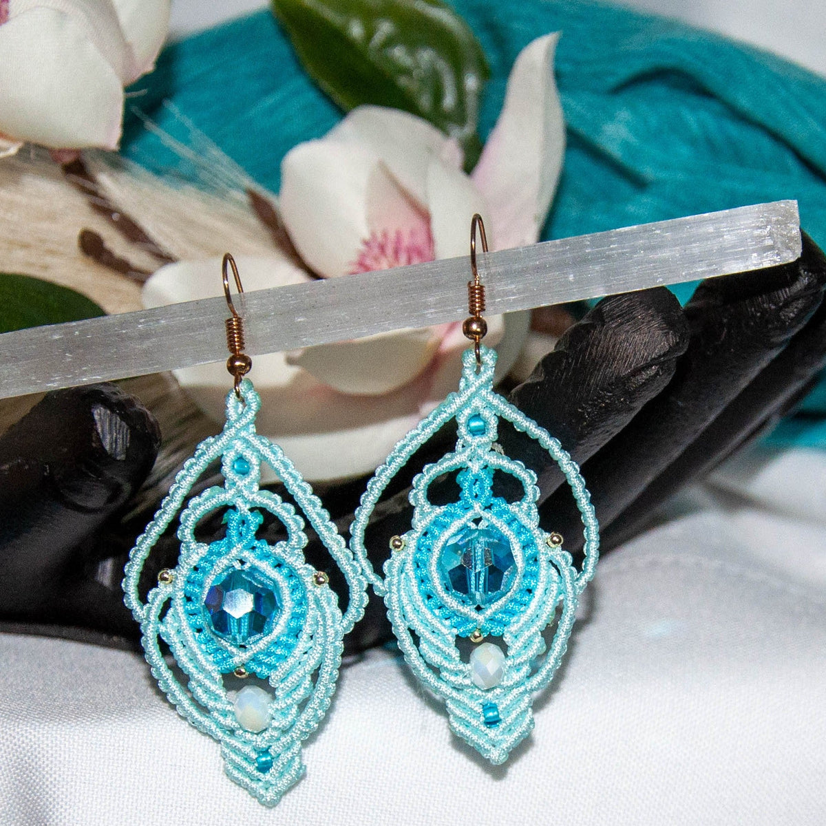 Handmade aqua two-tone lace macrame peacock earrings with faceted aqua glass bead, hanging from selenite wand with magnolia blossoms