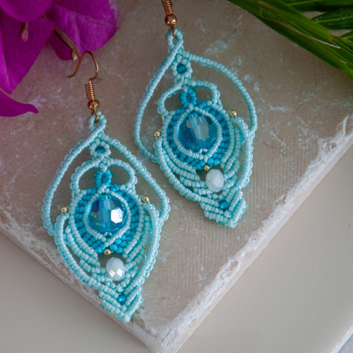 Pair of handmade aqua lace macrame peacock earrings with faceted glass and white rondelle, displayed on travertine tile with bougainvillea and tropical palm