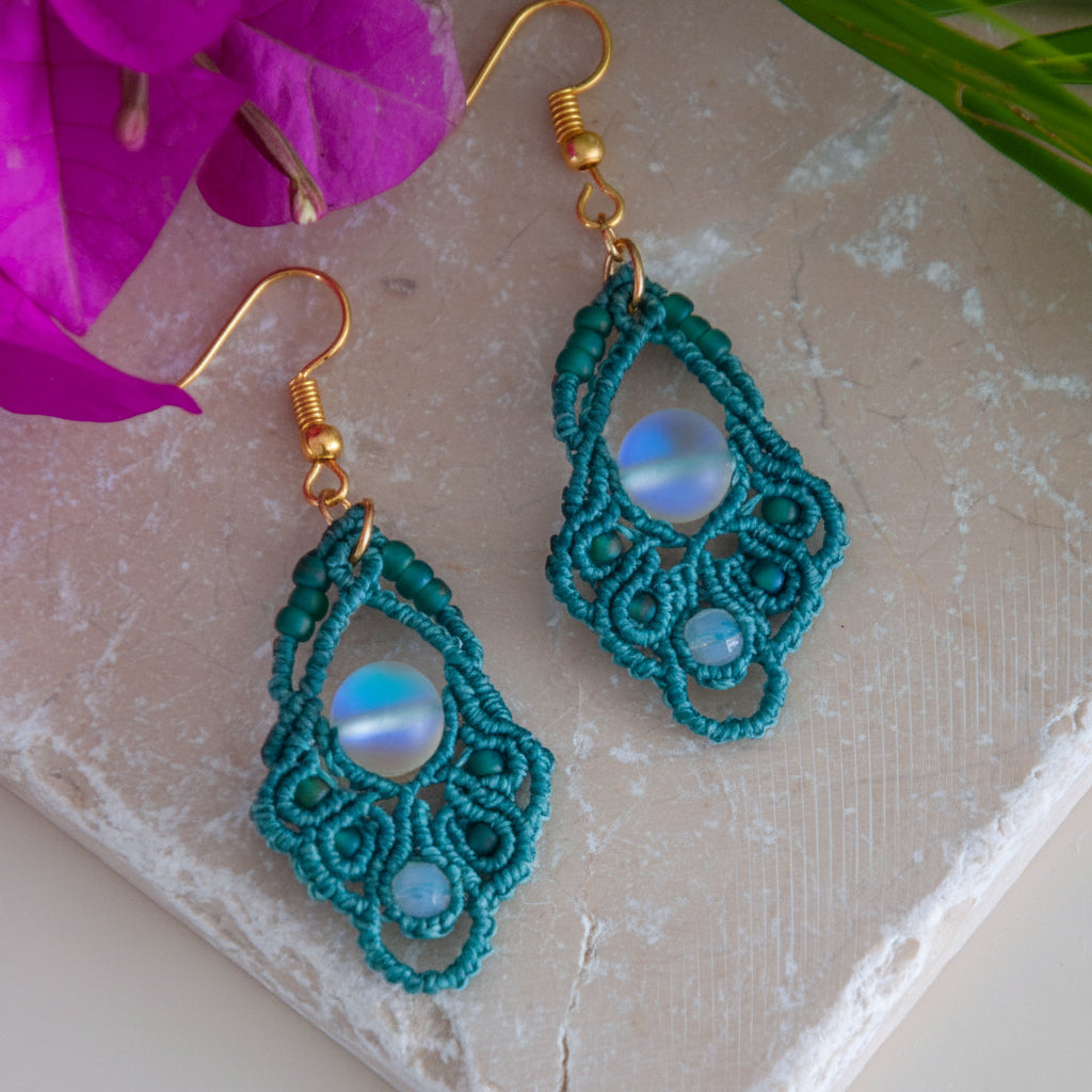 Handmade dark teal macrame diamond drop earrings with faceted opalite bead accents, teal seed beads, an iridescent frosted mermaid glass center bead, and gold-tone fish hook wires, laid on a travertine tile with magenta bougainvillea and tropical palm leaves.
