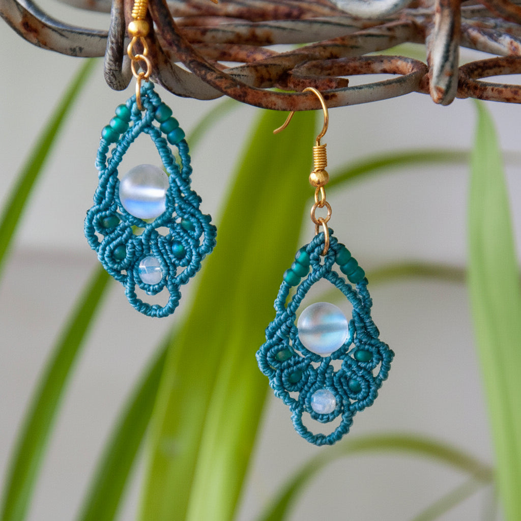 Handmade dark teal macrame diamond drop earrings with faceted opalite bead accents, teal seed beads, an iridescent frosted mermaid glass center bead, and gold-tone fish hook wires, hanging from a rustic metal stand with tropical green leaves in the background.