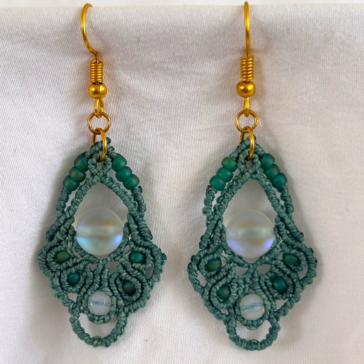 Pair of handmade dark teal macrame diamond drop earrings with 8mm iridescent frosted mermaid glass center bead, 4mm faceted opalite accents, teal seed beads, and gold-tone fish hook wires, displayed on white fabric background.
