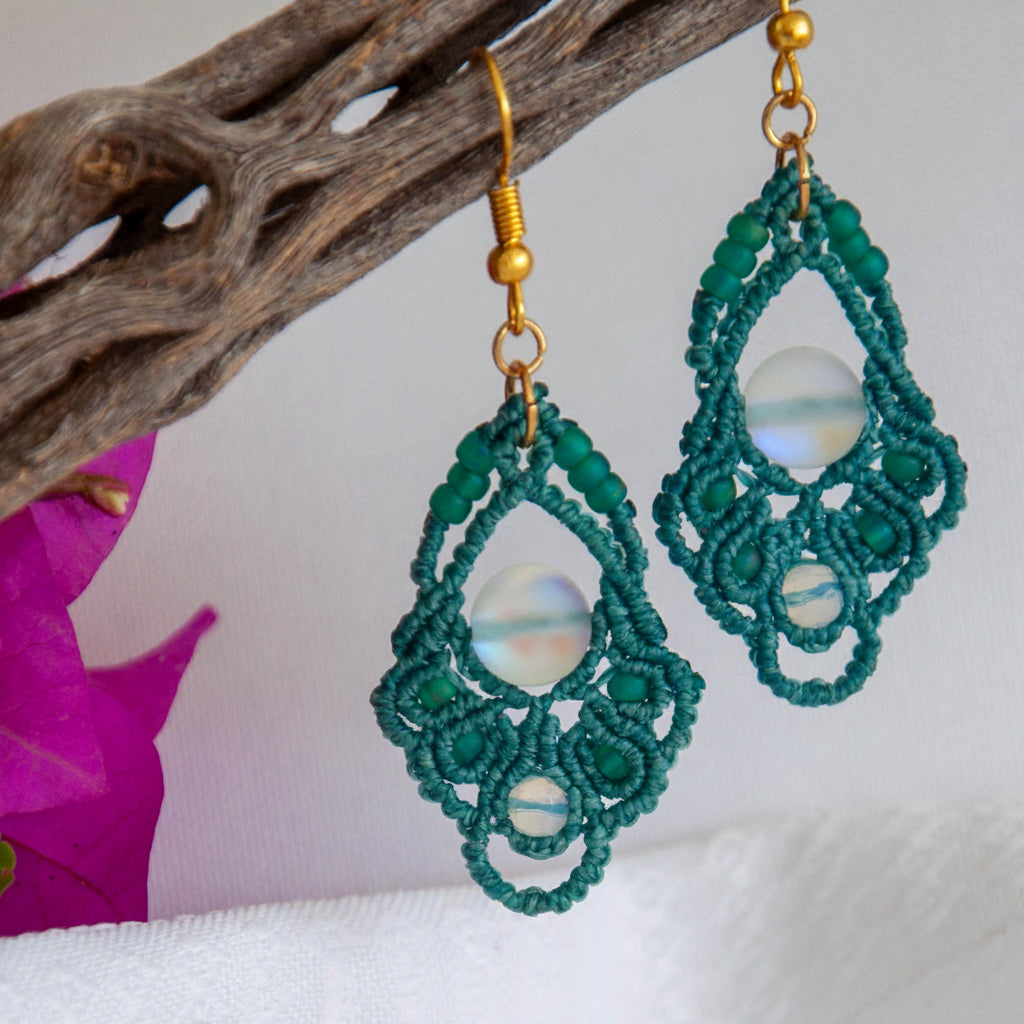 Handmade dark teal macrame diamond drop earrings with faceted opalite bead accents, teal seed beads, an iridescent frosted mermaid glass center bead, and gold-tone fish hook wires, hanging from a driftwood branch with magenta bougainvillea against a white background.