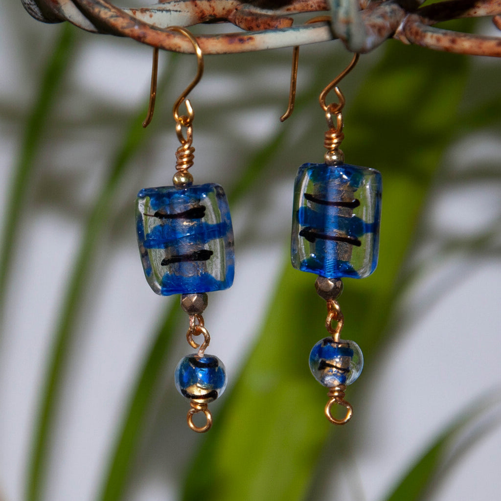 Handmade cobalt blue lampwork glass drop earrings with gold foil accents, coordinating round lampwork bead drops, and custom brass fish hook wires, hanging from a rustic metal stand with tropical green leaves.