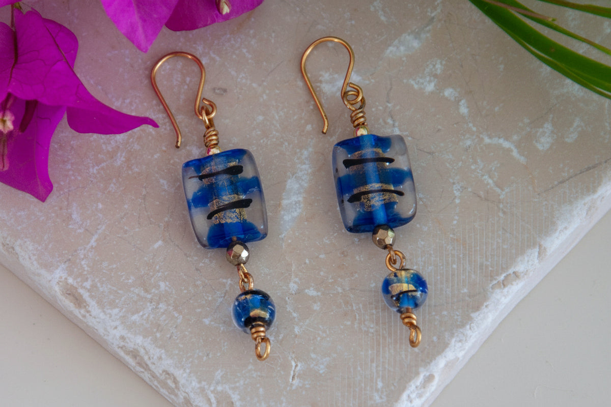 Handmade cobalt blue lampwork glass drop earrings with gold foil accents, coordinating round lampwork bead drops, and custom brass fish hook wires, laid flat on a natural stone surface with magenta bougainvillea flowers.