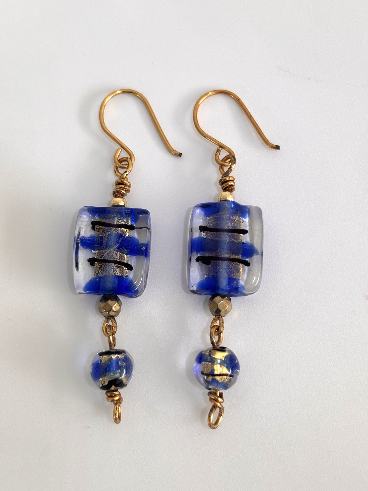 Pair of handmade cobalt blue lampwork glass drop earrings with gold foil detail, black brushstroke accents, faceted brass spacer beads, and 8mm coordinating round lampwork bead drops on custom brass fish hook wires, displayed on white background.