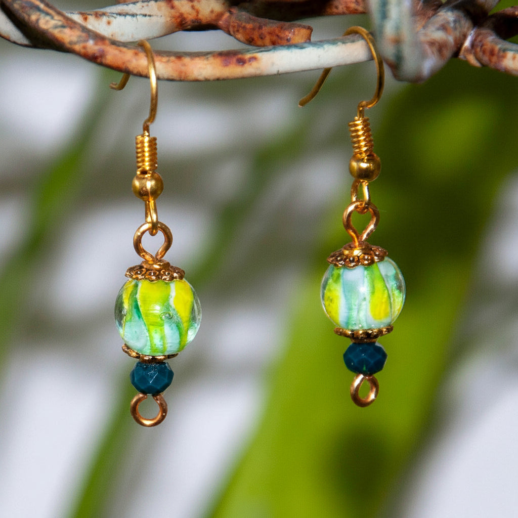 Handmade tropical swirl lampwork glass drop earrings with 8mm green, yellow, and white lampwork bead, gold-tone daisy spacer beads, and 3mm dark green faceted crystal rondelle, hanging from a rustic branch with blurred green foliage background.
