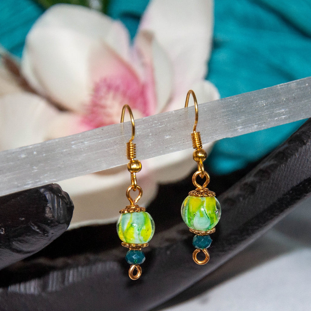 Handmade tropical swirl lampwork glass drop earrings with 8mm green, yellow, and white lampwork bead, gold-tone daisy spacer beads, and 3mm dark green faceted crystal rondelle, displayed on black slate with a selenite wand, white magnolia flower, and teal backdrop.