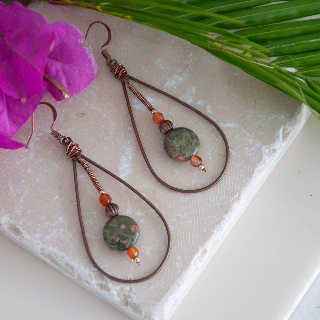 Pair of handmade copper teardrop earrings with unakite disc and carnelian beads, flat lay on travertine stone with bougainvillea and palm frond